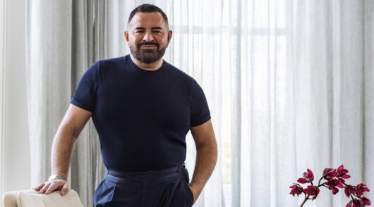 Mid shot of male Australian designer Steven Khalil in navy shirt and pants against neutral background in styled living room.