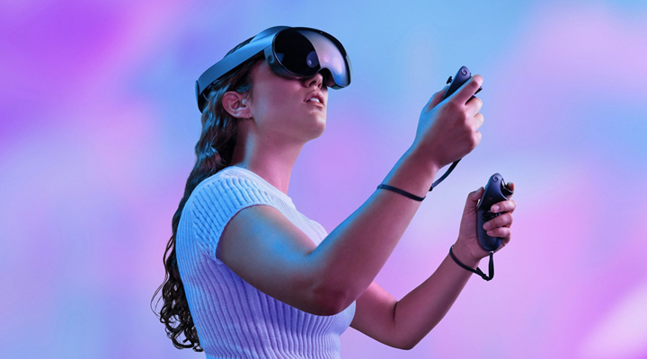 A young woman wearing a virtual reality headset and holding haptic handsets standing in front of a purple and blue background.