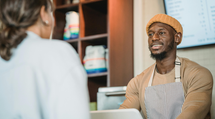 A Black man wearing an apron standing behind a register taking an order from a woman