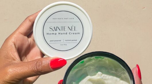 An image of a woman’s hand, with bright red nails, holding a Saint Nèl hand cream.