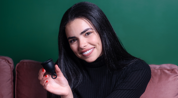Viv for your V founder Katie Diasti sitting against a dark green background holding a reusable menstrual cup.