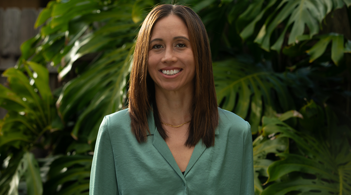a headshot of aloha collection's vp of supply chain maile english wearing a green shirt standing in front of green plants