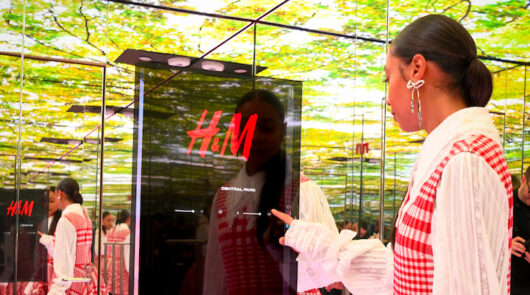 A young woman standing in front of a digital H&M display in a mirrored room.