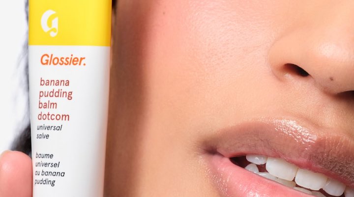 A close up of a female model holding a banana pudding flavored Glossier Balm Dotcom.