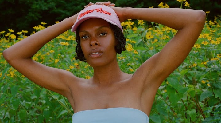 A young female model standing in a field of flowers wearing a blue strapless top and a pink Outdoor Voices baseball cap.