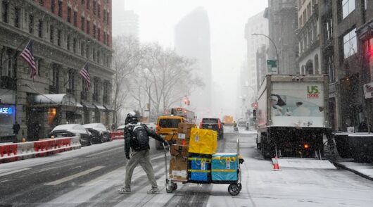 An Amazon delivery worker pulls their cart across the street during snowfall