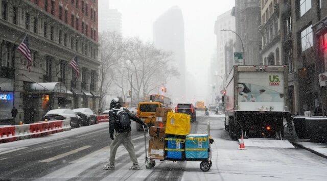 An Amazon delivery worker pulls their cart across the street during snowfall