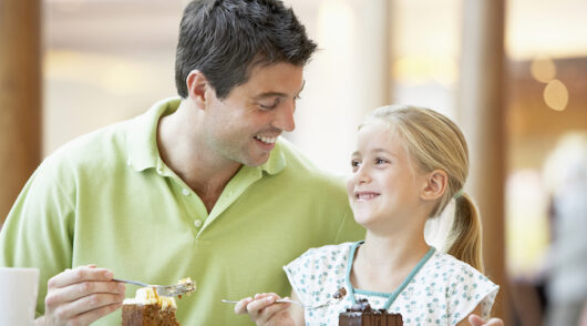 A father and daughter duo looking at one another as they sit at a restaurant eating cake.