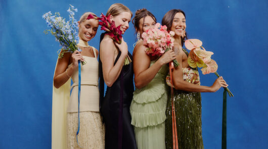 Four women standing side-by-side wearing formal wear dresses against a blue background.