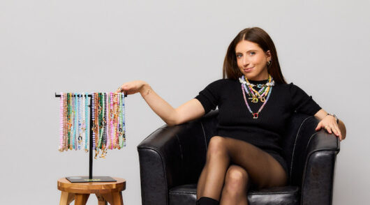 Spicy Dan founder Danielle Meyer holding up several necklaces and sitting in front of a gray background.
