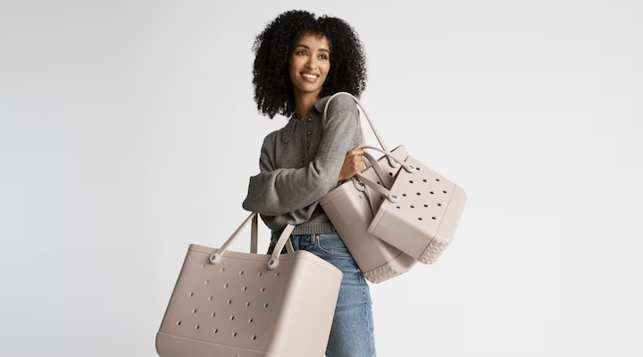 A female model wearing a grey sweater, jeans and sporting several light tan Bogg bags.
