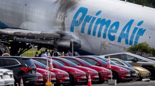 Tesla cars are parked in a lot next to where a crashed Amazon Prime Air cargo plane came to rest