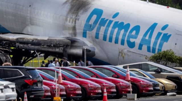 Tesla cars are parked in a lot next to where a crashed Amazon Prime Air cargo plane came to rest