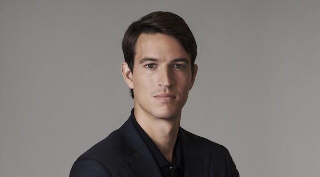 LVMH heir Alexandre Arnault standing in front of a grey background in a black shirt.
